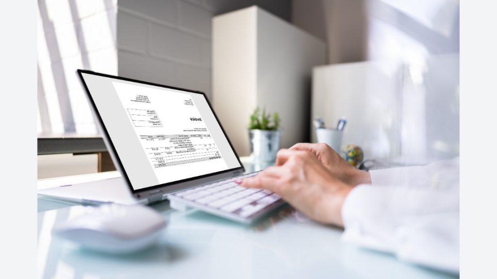 A person sits at a desk with a laptop open to an invoice.